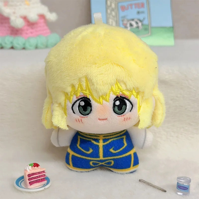 A plush doll with yellow hair and blue eyes, wearing a blue and yellow outfit, displayed on a white surface with small accessories including a miniature cake, a pen, and a small bottle.